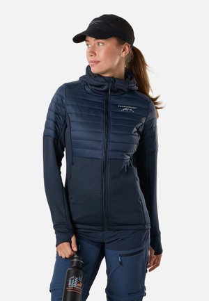 Kobieta w granatowej pikowanej kurtce i czapce trzyma czarną butelkę na wodę z logo outdoor, patrząc w bok na jednolitym tle.