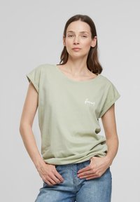 T-shirt en coton vert clair avec une coupe décontractée, des manches retroussées et le texte "forêt" en blanc sur le devant, associé à un jean en denim bleu.