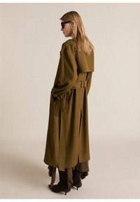 Abrigo largo verde oliva con un diseño estructurado, detalle de cinturón, mangas anchas y un pliegue en la parte posterior; combinado con capas marrón oscuro y botas hasta la rodilla.