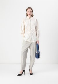 Camicia beige con ricami floreali, pantaloni grigi a pieghe, tacchi neri alti e una borsa blu. Collo semplice e chiusura con bottoni sulla camicia.