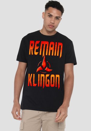 Young man wearing black T-shirt with red text "REMAIN KLINGON" and a red emblem, standing against a plain light background.