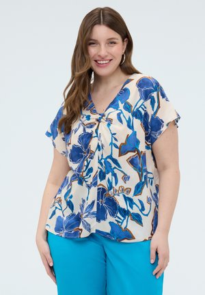 Femme souriante aux longs cheveux bruns portant des boucles d'oreilles en anneau, un chemisier crème à manches courtes avec un imprimé floral bleu, et un pantalon bleu vif sur un fond uni.