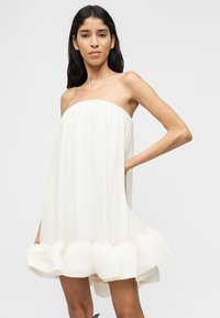 Γυναίκα που φοράει λευκό φόρεμα χωρίς τιράντες με βολάν στο τελείωμα, στέκεται με το ένα χέρι στη μέση μπροστά σε απλό φόντο.