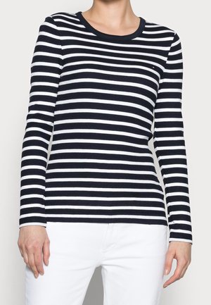 Femme portant un t-shirt à rayures horizontales noires et blanches à manches longues avec un pantalon blanc, debout devant un fond clair uni.
