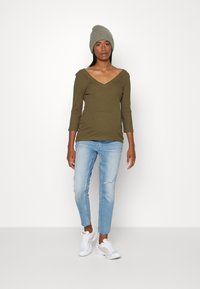 Top de manga longa em verde-oliva com nervuras e decote em V, combinado com jeans claros desgastados e ténis brancos. O modelo usa um gorro de malha cinza.