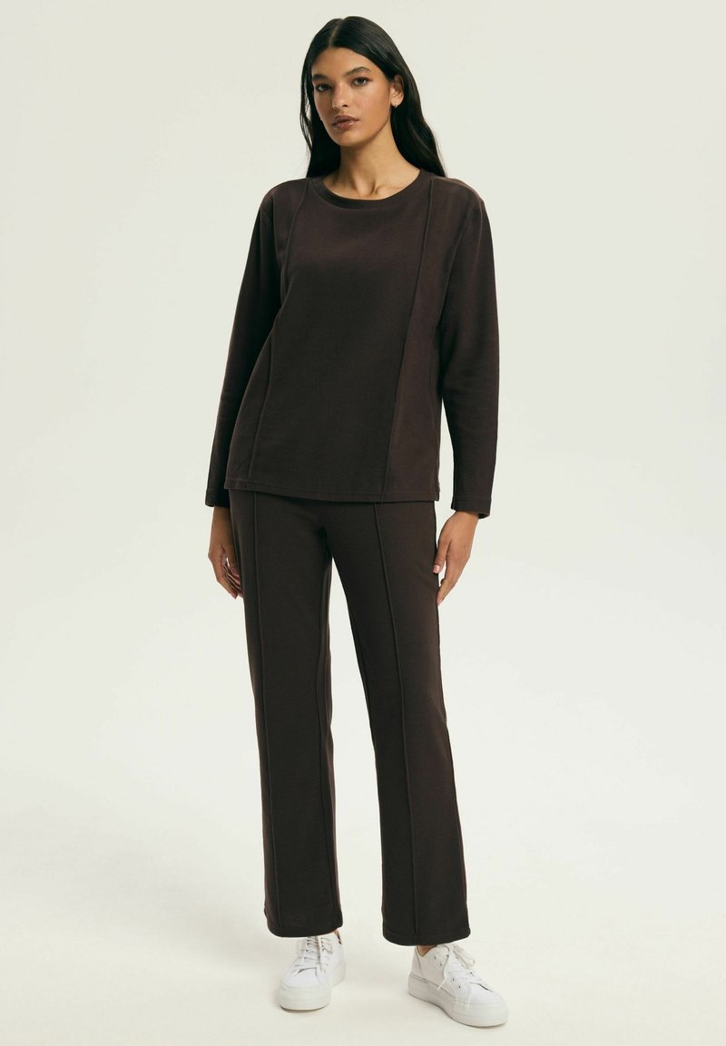 Brązowa bluzka z długim rękawem i zaokrąglonym dekoltem, zestawiona z pasującymi brązowymi spodniami. Oba elementy mają przeszywane szwy i wykonane są z miękkiego materiału.