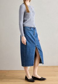 Šedý kardigan s dlouhým rukávem zapnutý vpředu na knoflíky, kombinovaný s modrou džínovou sukní po kolena s předním rozparkem, nošený s černými balerínami.