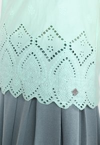 Mintgrön broderad topp med intrikata utskärningar av spetsmönster vid fållen, kombinerad med en mjuk grå plisserad kjol.