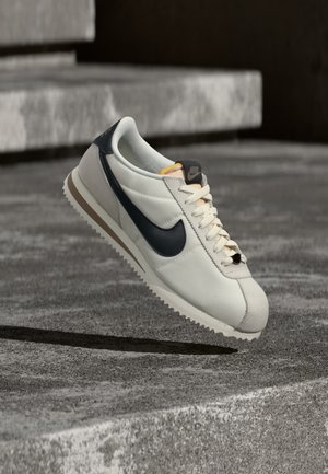 Hvid Nike Cortez-sneaker med sort swoosh-logo, beige sål og snørebånd, placeret over grå betontrapper og -jord.