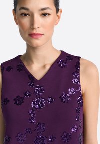 Fioletowy top bez rękawów z dekoltem w kształcie litery V, ozdobiony kwiatowymi wzorami z cekinów w różnych odcieniach fioletu, tworzącymi teksturę i połysk.