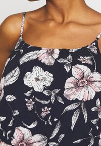Top à imprimé floral avec bretelles spaghetti, au fond marine, avec des fleurs roses et blanches, et une texture de tissu lisse. Détail de décolleté accentué.