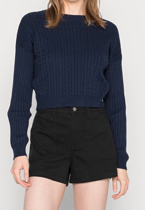 Vrouw draagt een cropped kabelgebreide trui met lange mouwen in marineblauw en een zwarte high-waisted denim short, staand tegen een effen achtergrond.