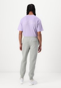 Μπλουζάκι λεβάντας με λευκό κείμενο στην πλάτη, σε συνδυασμό με ανοιχτό γκρι φόρμα και λευκά αθλητικά παπούτσια. Το σύνολο έχει χαλαρή γραμμή.