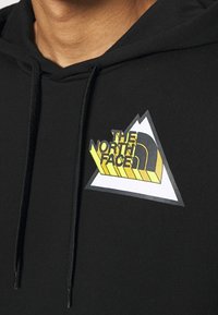 Felpa con cappuccio nera con un logo sul petto anteriore in giallo e grigio, raffigurante montagne e il testo "The North Face" in uno stile di carattere moderno.