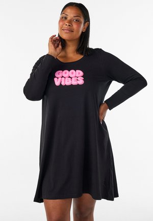 Frau trägt ein schwarzes langärmeliges Kleid mit pinkem Text "GOOD VIBES", posiert mit einer Hand am Hals und der anderen an der Hüfte.