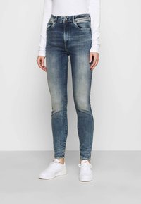 Jeans ajustados en denim azul desgastado, diseño de cintura alta con cinco bolsillos y un sutil desgastado. Combinados con zapatillas blancas.