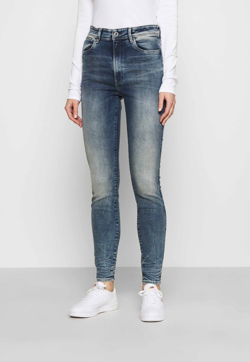 Jeans ajustados en denim azul desgastado, diseño de cintura alta con cinco bolsillos y un sutil desgastado. Combinados con zapatillas blancas.