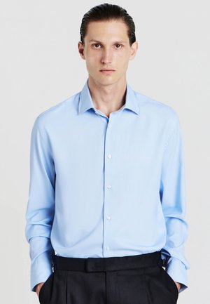 Camicia button-up azzurro chiaro con maniche lunghe e colletto a punta, caratterizzata da una texture liscia e un sottile motivo a righe. Abbinata a pantaloni scuri.