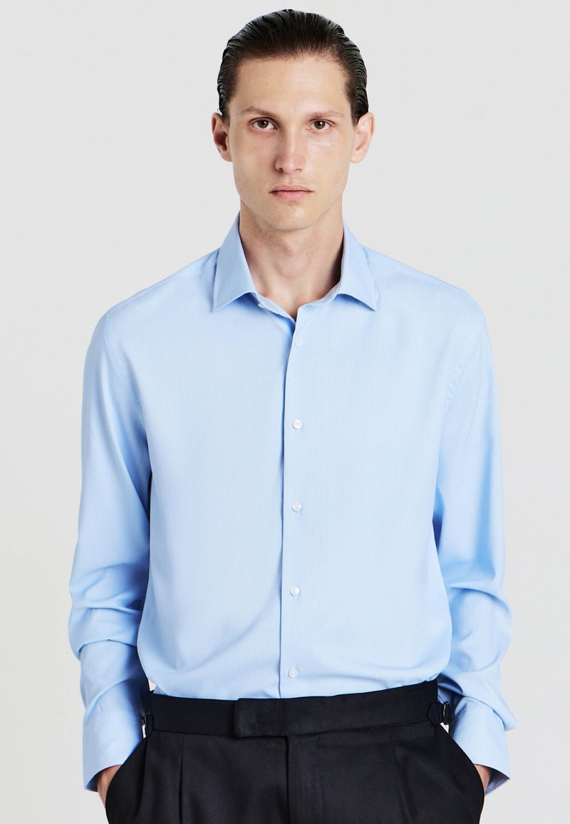 Camicia button-up azzurro chiaro con maniche lunghe e colletto a punta, caratterizzata da una texture liscia e un sottile motivo a righe. Abbinata a pantaloni scuri.