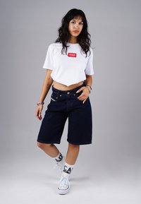 Young woman with long dark hair wearing a cropped white t-shirt, dark denim shorts, white socks with text, and white sneakers, standing on gray background.