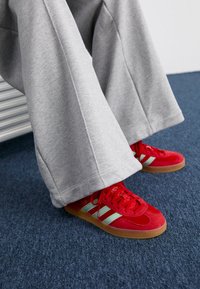 Červené sportovní boty se světle modrými pruhy a gumovými podrážkami, v kombinaci se šedými volnými tepláky na modrém koberci.