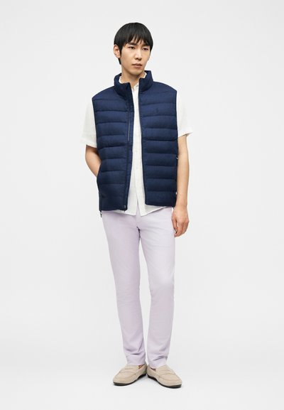 Άνδρας που στέκεται φορώντας ένα σκούρο μπλε καπιτονέ γιλέκο πάνω από λευκό κοντομάνικο πουκάμισο, ανοιχτόχρωμο μωβ παντελόνι και μπεζ παπούτσια χωρίς κορδόνια σε λευκό φόντο.