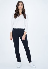Top bianco leggero a maniche lunghe con scollo a V, abbinato a pantaloni neri aderenti e sneakers bianche. Design semplice, senza pattern o dettagli visibili.
