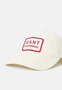 Kapa od kremaste tkanine s crvenim vezom s natpisom "GANT SHIRTMAKERS". Zakrivljeni vizir i podesiva traka na poleđini.