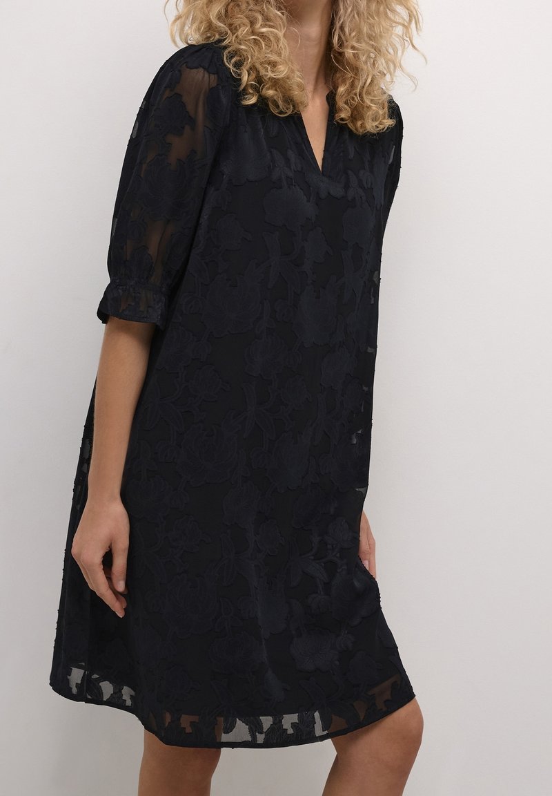 Personne aux cheveux bouclés portant une robe ample en dentelle noire à motifs floraux avec des manches arrivant aux coudes, sur un fond clair uni.