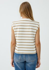 Top bianco senza maniche con sottili strisce dorate, caratterizzato da una vestibilità ampia e un collo rotondo. Indossato con jeans di denim blu a vita alta.
