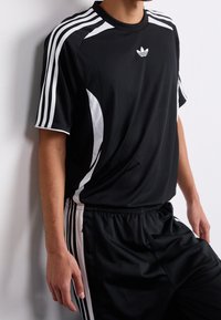 Persona che indossa un abbigliamento sportivo Adidas nero con strisce bianche sulle maniche e sui pantaloni, appoggiata a un muro bianco.