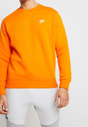 Homme portant un sweat Nike orange vif et un pantalon Nike gris clair avec des logos sur la poitrine et la cuisse, debout devant un fond uni.