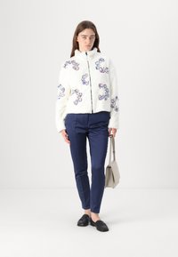 Weiße Kunstpelzjacke mit buntem Blumenstickerei, Reißverschluss und hohem Kragen, kombiniert mit navy-farbenen, engen Hosen und schwarzen Slippern.