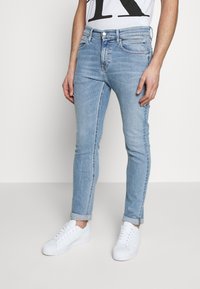 Jeans de denim azul claro con corte slim, que cuentan con cinco bolsillos y un leve desvanecimiento. Combinados con zapatillas deportivas blancas, con los puños enrollados visibles.