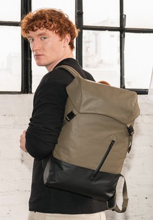Khaki and black backpack with a structured design, featuring a top flap, side zipper pocket, and adjustable straps. Smooth texture and matte finish.