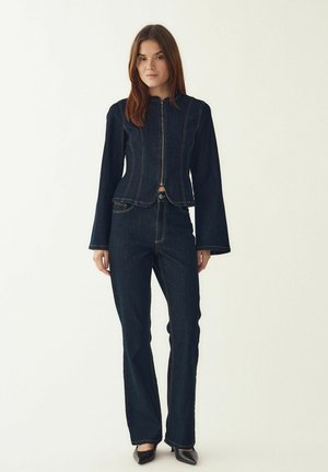 Chaqueta de mezclilla oscura y vaqueros acampanados a juego, con cremallera frontal, pespuntes decorativos y diseño corto. Calzado negro.