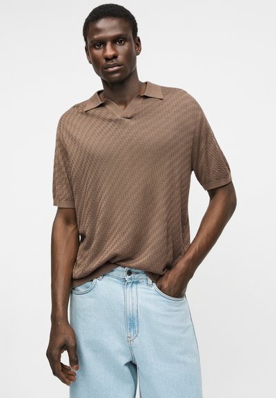 Homme portant un polo marron texturé à manches courtes, rentré dans un jean bleu clair, avec une main dans la poche.