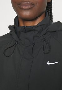 Černá bunda na zip s kapucí a stahovacími šňůrkami. Na levé straně hrudi je viditelná bílá logo Nike ve tvaru křídel.
