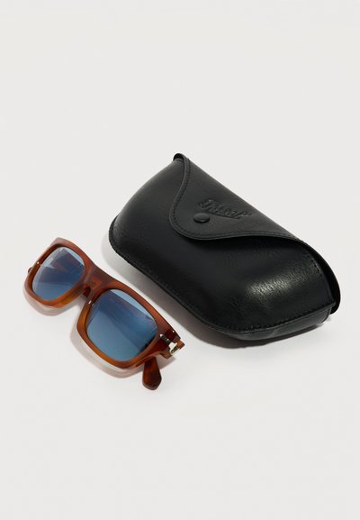 Καφέ ορθογώνια γυαλιά ηλίου με μπλε φακούς δίπλα σε μια μαύρη δερμάτινη θήκη με κούμπωμα κουμπί, σε λευκό φόντο.