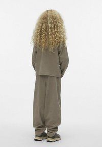 Brązowy prążkowany sweter i dopasowane luźne spodnie. Ma długie kręcone blond włosy i neutralne kolory sportowych butów. Prosty, czysty design.