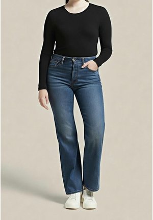 Persoon draagt een zwart topje met lange mouwen, blauwe jeans met rechte pijpen, witte schoenen, met één hand in de zak, staand tegen een effen achtergrond.