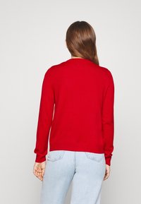 Maglione rosso a maglia con scollatura rotonda e maniche lunghe. Presenta polsini e orlo a costine. Abbinato a jeans azzurri chiari.