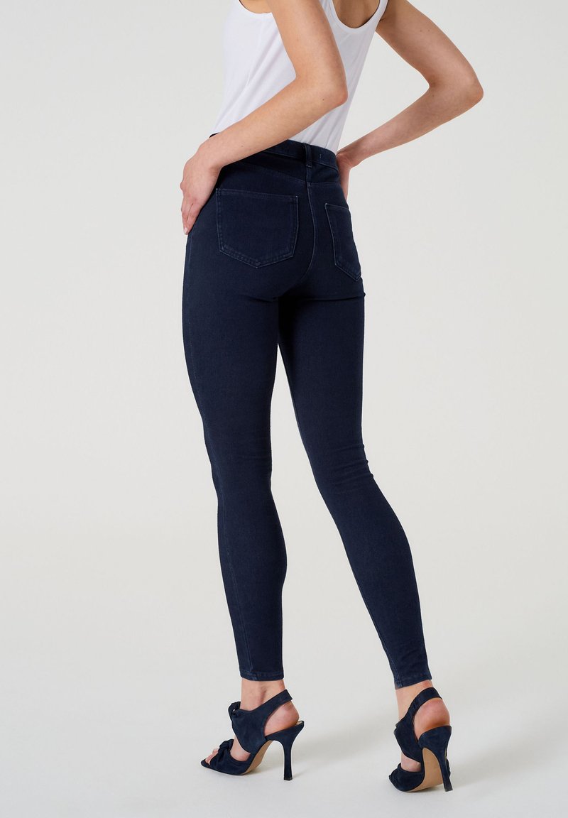 Tmavě modré skinny džíny s pěti kapsami, vysokým pasem a hladkým povrchem. Noseny s černými sandály na vysokém podpatku.