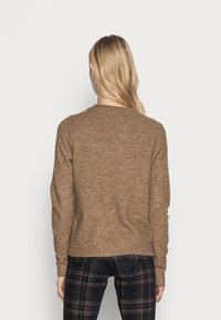 Blonde Frau von hinten gesehen, die einen braunen Strickpullover und schwarze Karohosen trägt, vor einem schlichten hellen Hintergrund.