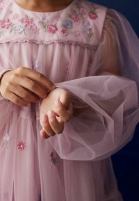 Hellrosa Kleid mit floralem Stickereimuster, durchsichtigen Puffärmeln und einem geschmeidigen Halsauschnitt. Hände, die den Ärmelsaum anpassen.