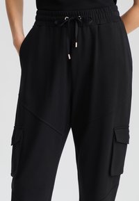 Pantaloni jogger neri con vita elastica, cordoncino regolabile e due tasche laterali cargo; tessuto liscio e leggero con dettagli sulle cuciture.
