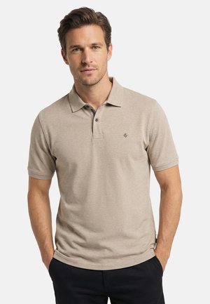 Man wearing beige short-sleeve polo shirt with collar and dark buttons, standing with hands in pockets against plain background.