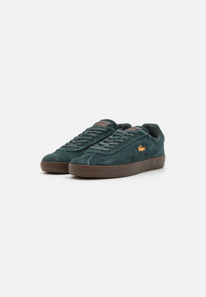 Zapatillas de ante verde con suela de goma marrón, acabado texturizado y cordones. Presentan un pequeño parche de logo naranja en la lengüeta y en el costado.