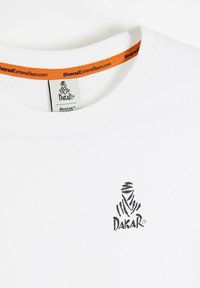 Biała bawełniana koszulka z czarnym logo Dakar na klatce piersiowej i pomarańczową metką na wewnętrznej części dekoltu, z krótkimi rękawami i okrągłym dekoltem.