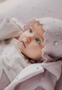 Bébé portant un doux bonnet tricoté rose avec des bords festonnés, une robe claire ornée de motifs floraux et de détails en nœud, blotti dans un tissu douillet.